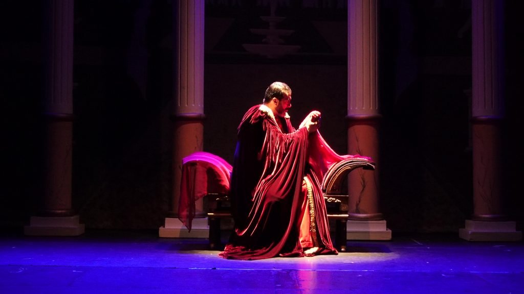 Escena teatral de La Voz Poderosa con un personaje inclinado sobre un manto rojo